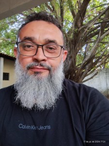 A imagem mostra um homem com uma longa barba grisalha e cabelo curto, usando óculos e uma camisa preta com o logotipo "Calvin Klein Jeans" estampado no peito. Ele está em um ambiente externo, com uma árvore grande e galhos densos ao fundo. A iluminação natural destaca o rosto e os detalhes da barba, criando uma atmosfera tranquila e natural. A expressão facial é neutra, transmitindo uma aparência calma e acolhedora. No canto inferior direito, há uma marcação de data e hora: "17 de jul. de 2024, 17:26."