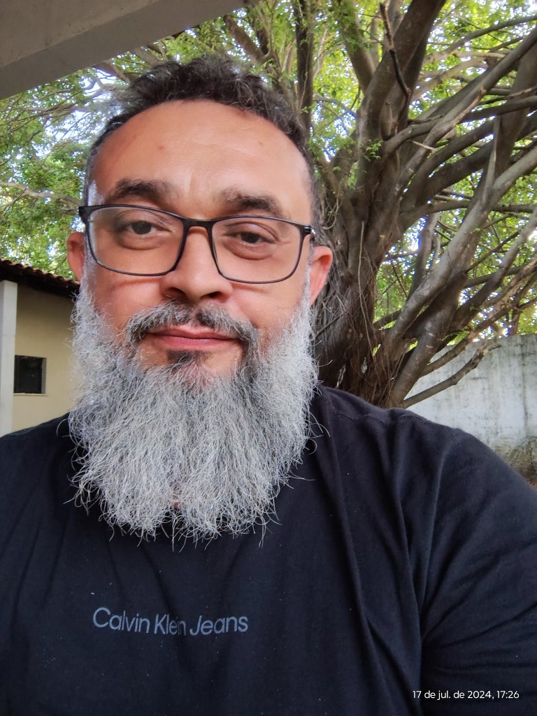 A imagem mostra um homem com uma longa barba grisalha e cabelo curto, usando óculos e uma camisa preta com o logotipo "Calvin Klein Jeans" estampado no peito. Ele está em um ambiente externo, com uma árvore grande e galhos densos ao fundo. A iluminação natural destaca o rosto e os detalhes da barba, criando uma atmosfera tranquila e natural. A expressão facial é neutra, transmitindo uma aparência calma e acolhedora. No canto inferior direito, há uma marcação de data e hora: "17 de jul. de 2024, 17:26."